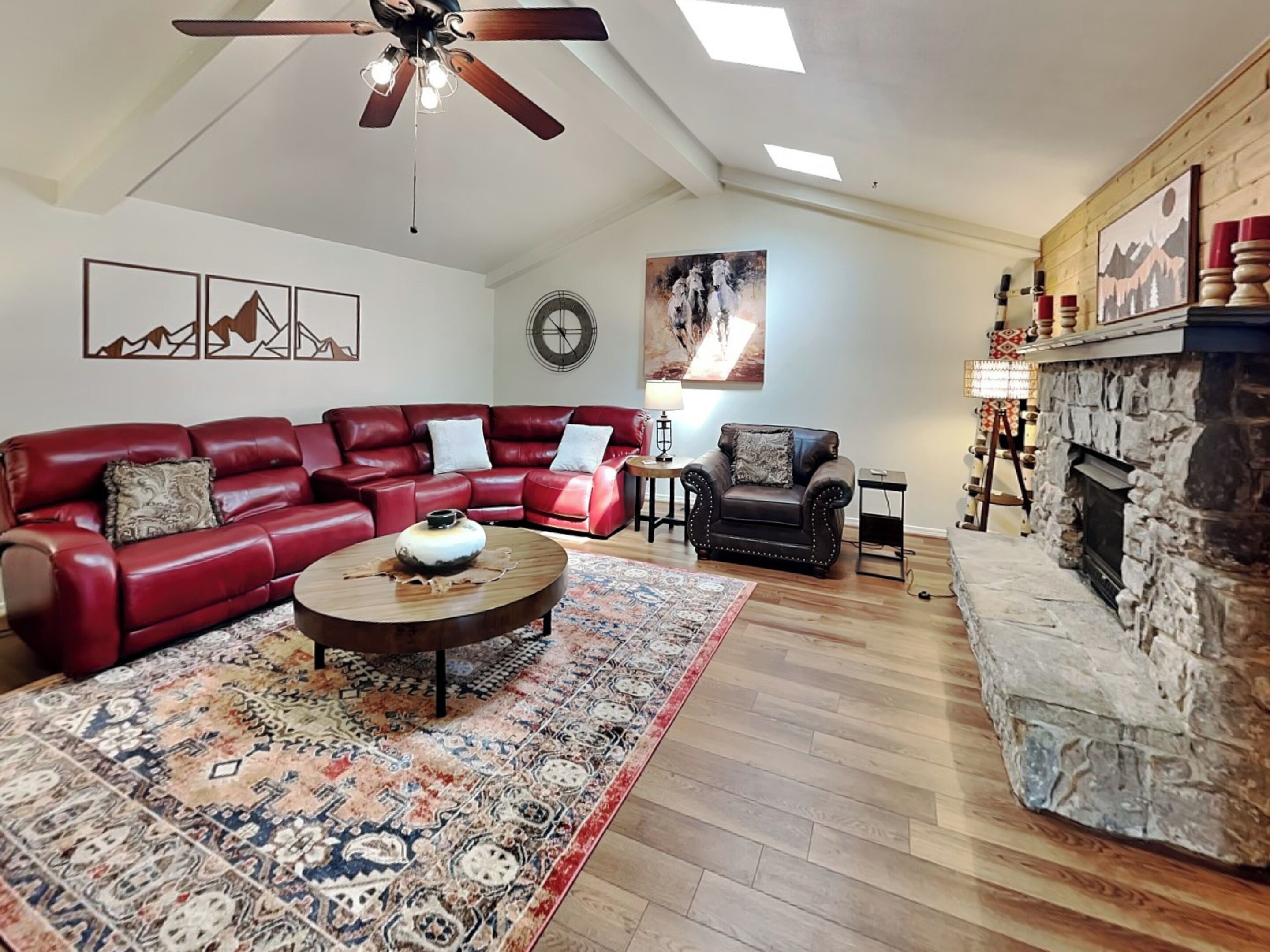 A spacious living room with a red leather sectional sofa, a black armchair, and a round coffee table. A stone fireplace is visible on the right, and a ceiling fan hangs above.