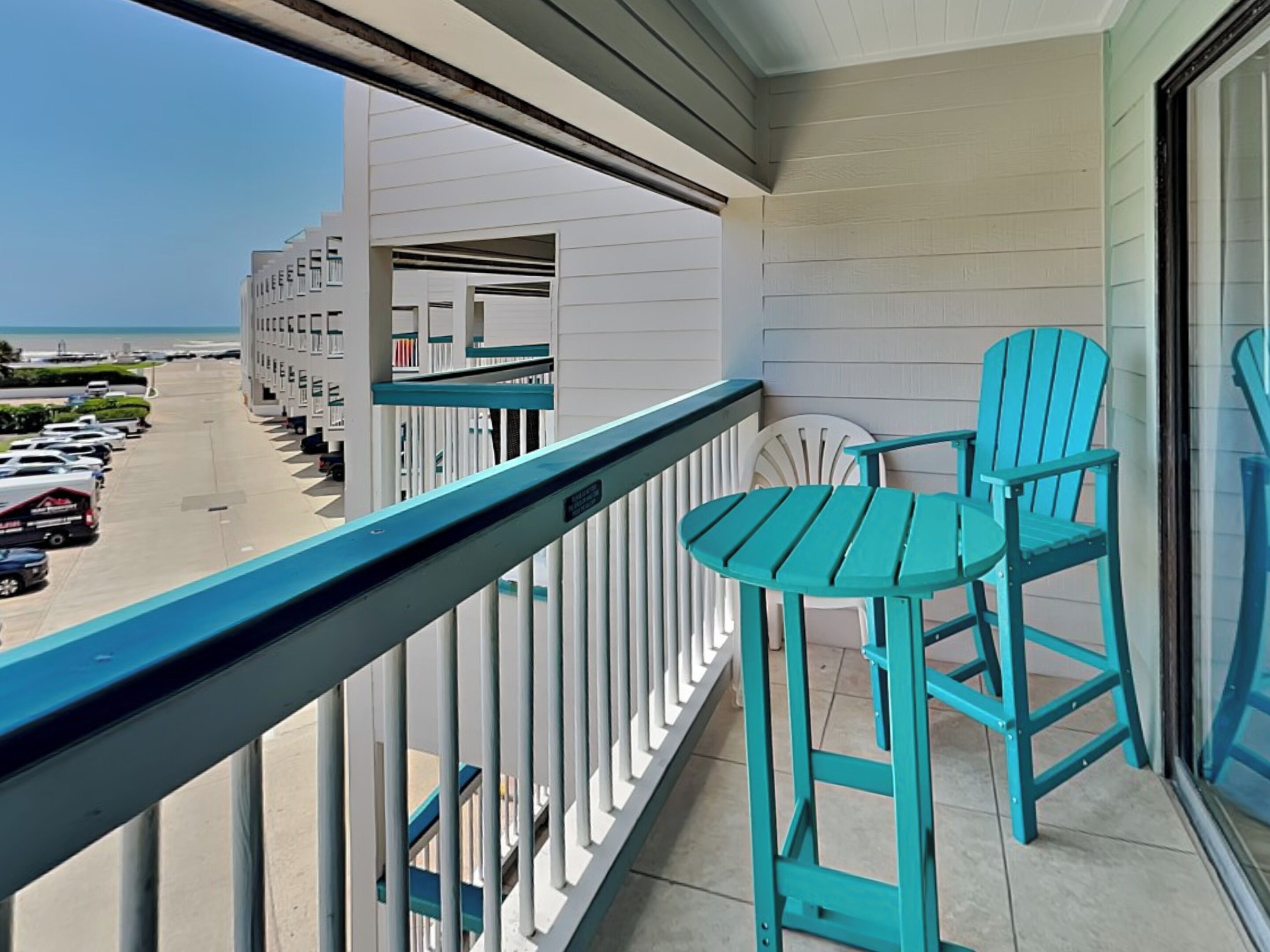 A balcony with turquoise chairs and a view of a parking lot and buildings.