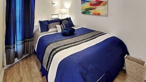 Un dormitorio con una cama grande, una mesita de noche, una lámpara, un cuadro en la pared y una ventana con cortinas.