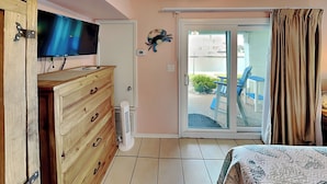 Un dormitorio con televisor, cómoda de madera y puerta corrediza de vidrio que da a un balcón con mobiliario exterior.