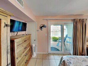 A bedroom with a TV, wooden dresser, and a sliding glass door leading to a balcony with outdoor furniture. - Mermaids Den by Miss Kitty’s (Rockport)