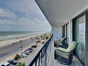 Un balcon avec deux fauteuils en osier, une vue sur la plage et des voitures garées le long de la route.