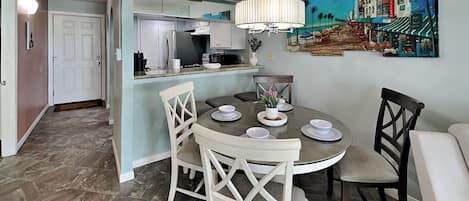 Uma sala de jantar com uma mesa redonda, cadeiras e um lustre. Uma cozinha aparece ao fundo, com armários e uma geladeira.
