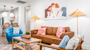 Una sala moderna con un sofá azul, un sofá marrón y una mesa de centro de madera con revistas. Hay dos lámparas de pie y un cuadro en la pared.