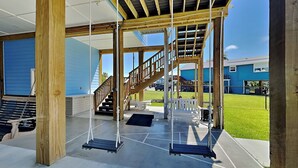 A covered outdoor area with a wooden swing set, a staircase, and a view of a blue building in the background. - Play N Hooky by Ryson Vacation Rentals (Galveston)