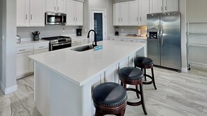 A modern kitchen with white cabinets, stainless steel appliances, a central island with stools, and a marble floor. - Play N Hooky by Ryson Vacation Rentals (Galveston)