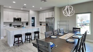A modern kitchen with white cabinets, a central island with stools, a dining area with a wooden table and chairs, and a large window providing a view of the outside. - Play N Hooky by Ryson Vacation Rentals (Galveston)