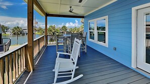 A covered deck with a view of a lake, wooden chairs, and a ceiling fan. - Play N Hooky by Ryson Vacation Rentals (Galveston)