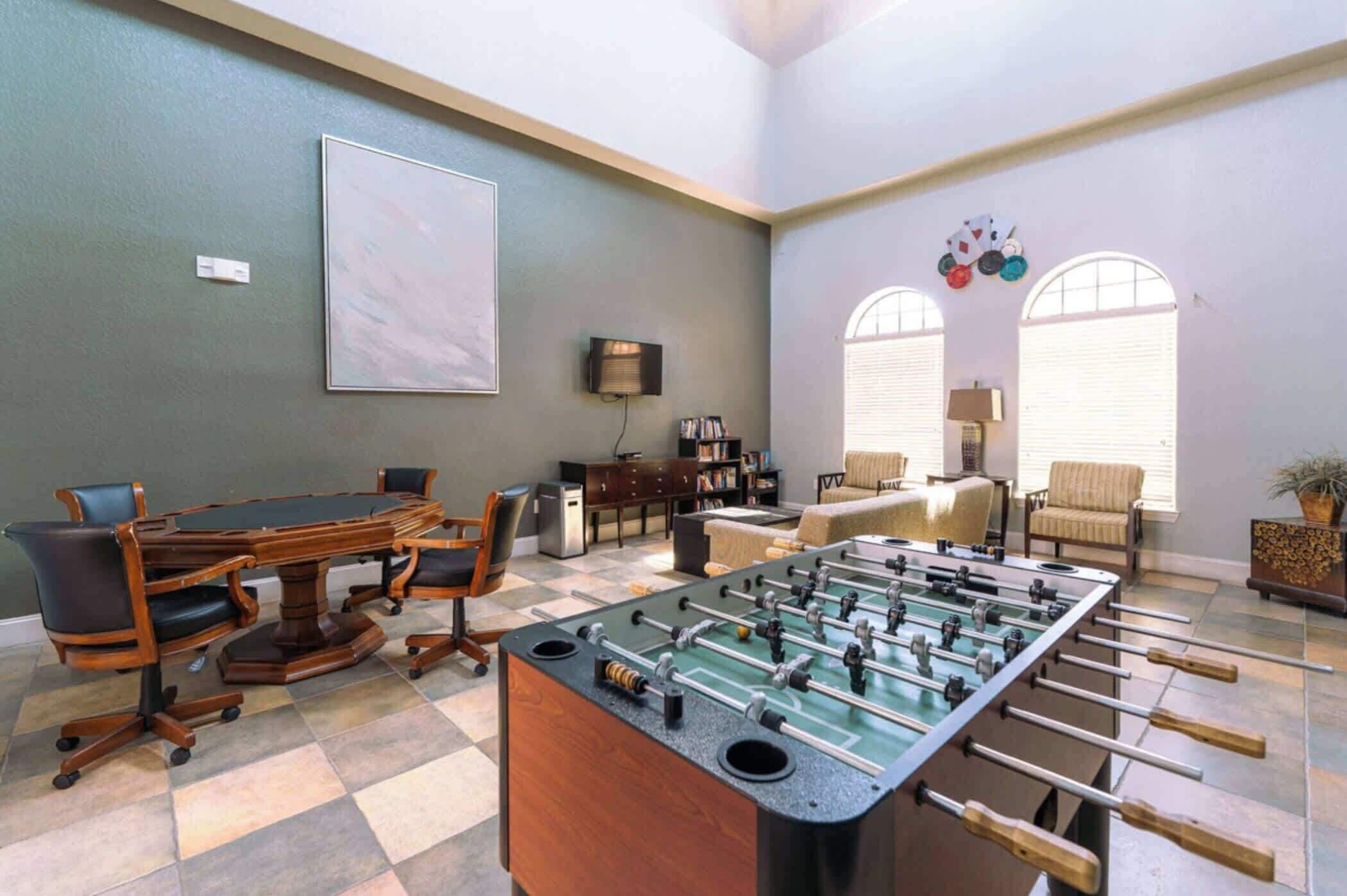 A room with a foosball table, a conference table, and a sofa. There are two windows with blinds, a bookshelf, and a lamp.