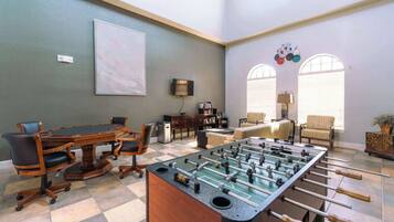 A room with a foosball table, a conference table, and a sofa. There are two windows with blinds, a bookshelf, and a lamp.