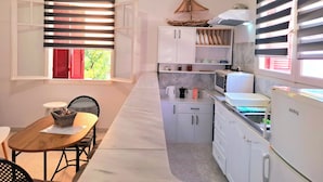 A compact kitchen with white cabinets, a microwave, a kettle, and a refrigerator. There is a dining area with a table and chairs, and a window with blinds. - Alegria Apartments (Paxos)