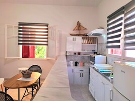 Cocina compacta con armarios blancos, microondas, hervidor y refrigerador. Hay una zona de comedor con mesa y sillas, y una ventana con persianas.