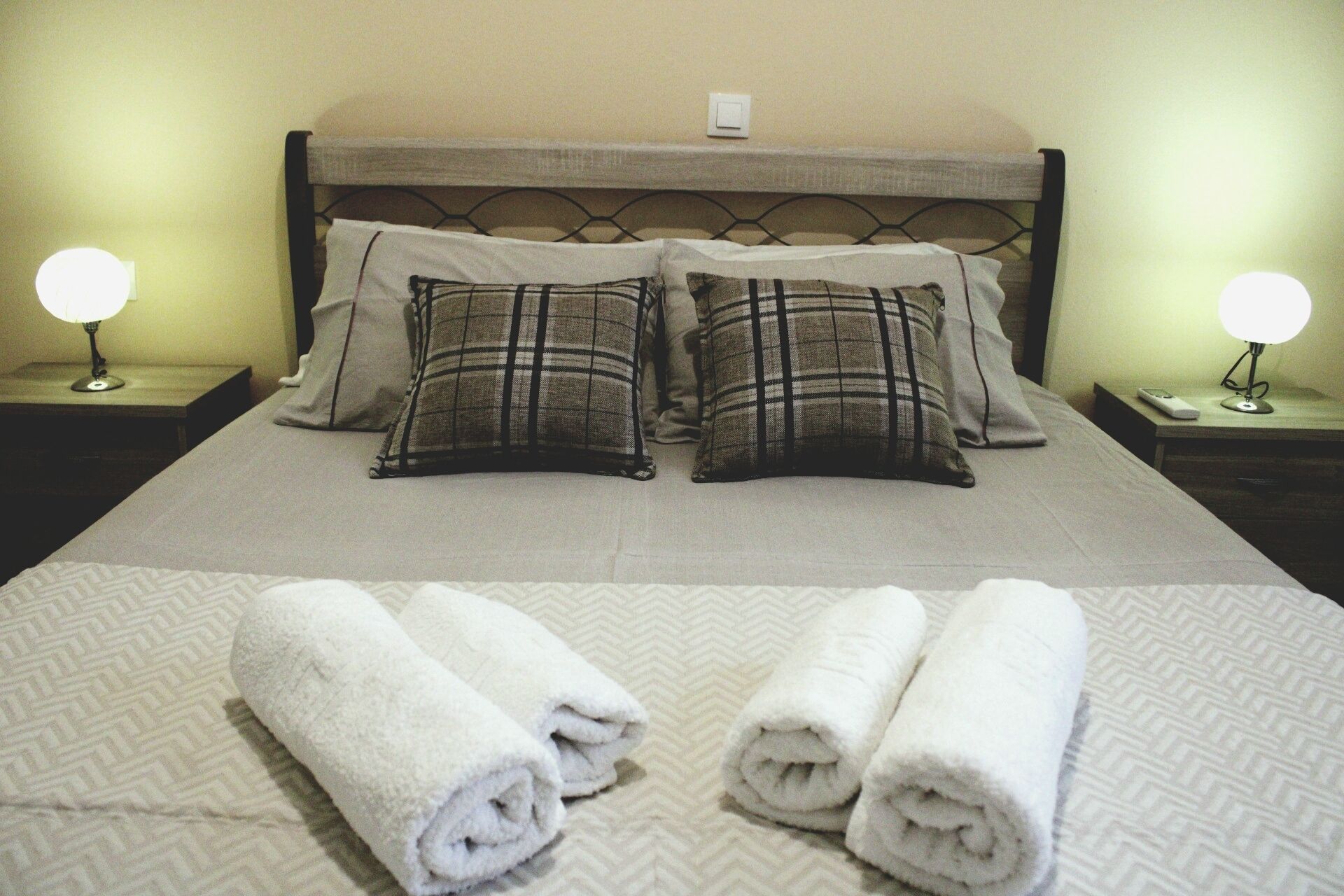 A neatly made bed with plaid pillows, two bedside lamps, and rolled towels on a patterned bedspread.