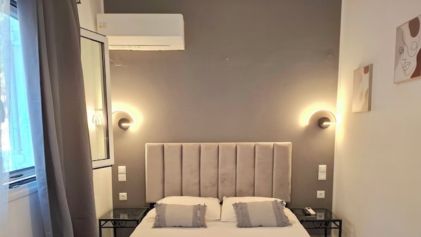 A hotel room with a bed, bedside tables, an air conditioning unit, and wall-mounted light fixtures. - Alegria Apartments (Paxos)