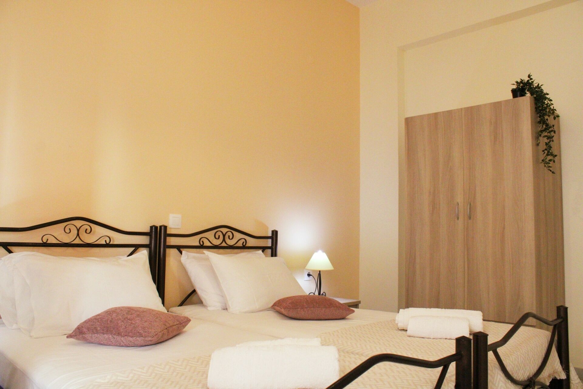 Una cama bien hecha con almohadas blancas y rosas, una lámpara de noche y un armario de madera en una habitación con paredes beige.