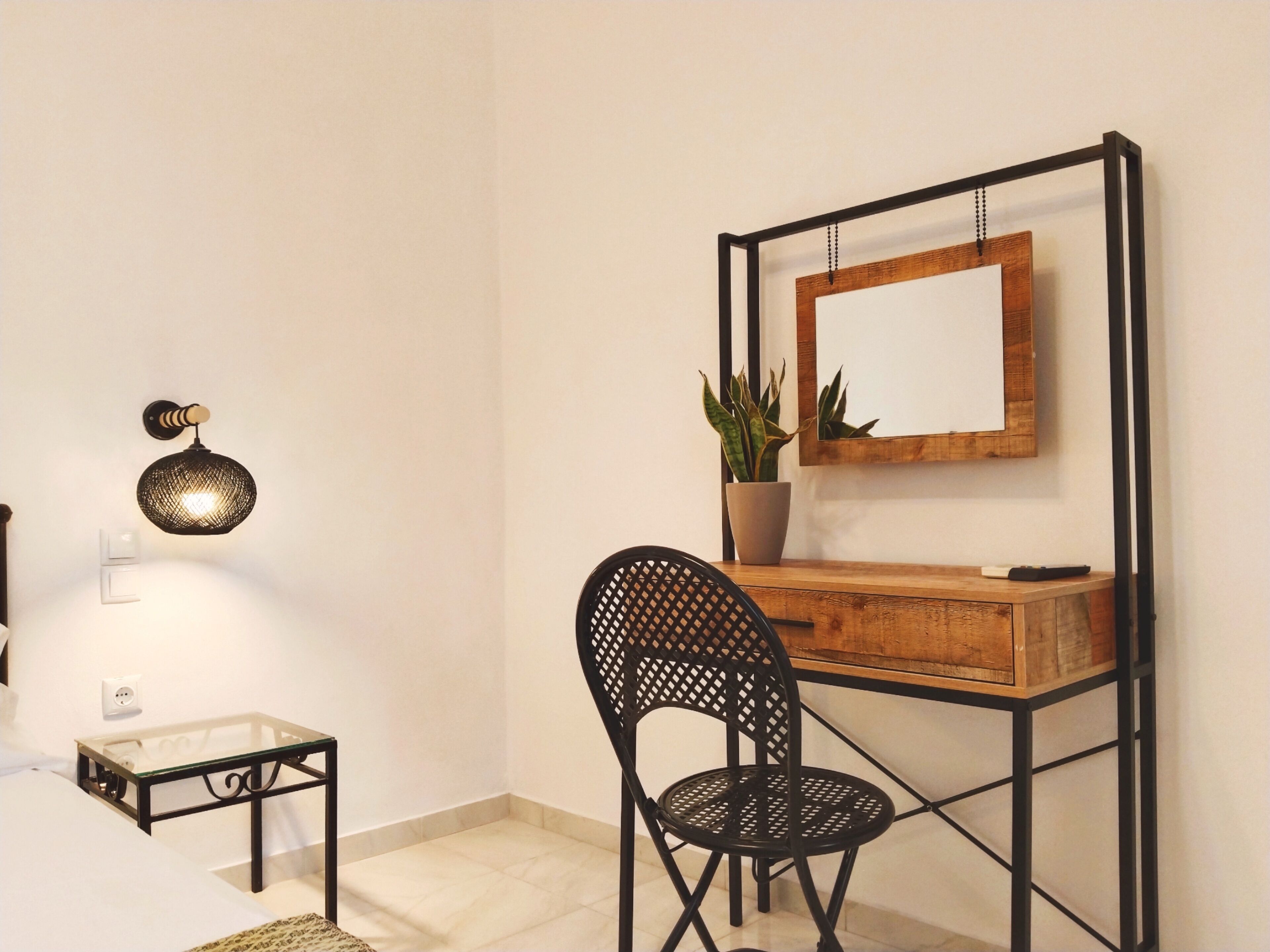 Quarto com escrivaninha de madeira, cadeira preta, planta em vaso, espelho fixado na parede e luminária na parede.