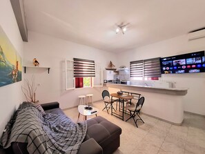 A compact living space with a sofa, a small dining table, and a kitchen area with a TV. - Alegria Apartments (Paxos)