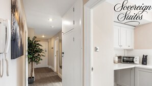 A modern kitchen with white cabinets, a microwave, and a knife block. A hallway with a potted plant and a door leading to another room is visible.