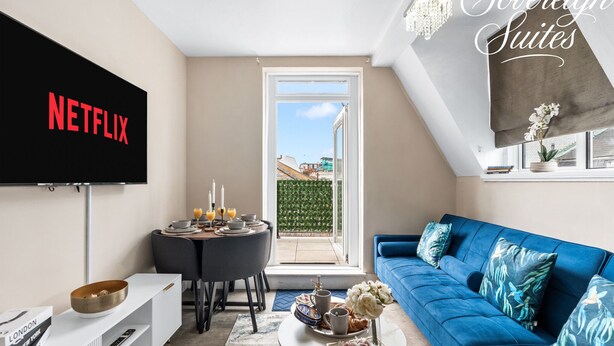 A living room with a TV, a blue sofa, a dining table, and a view of the outside.