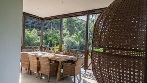 Un espace extérieur abrité, aménagé pour les repas, avec une grande table et des chaises en osier, de grandes fenêtres et une vue sur une colline boisée.