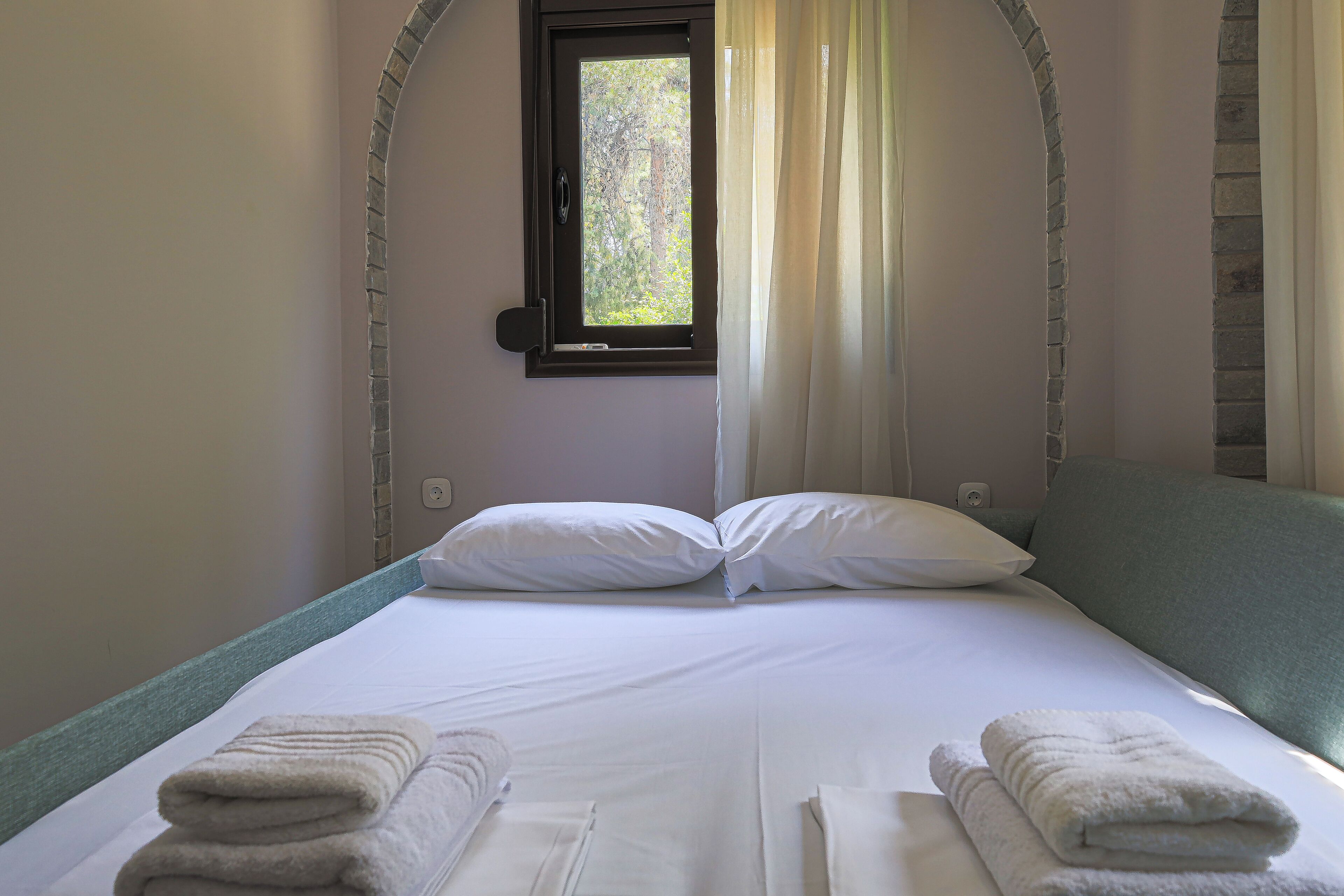 A neatly made bed with white linens and towels, a window with a view of trees, and a brick wall.