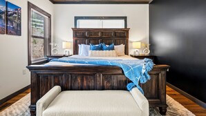 A bedroom with a large bed, wooden headboard, two bedside tables with lamps, a window with blinds, and a view of a mountain.