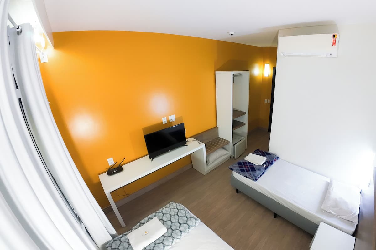 Ein kompaktes Hotelzimmer mit Bett, einem kleinen Schreibtisch mit Fernseher, einem Kleiderschrank und einer eingebauten Klimaanlage.