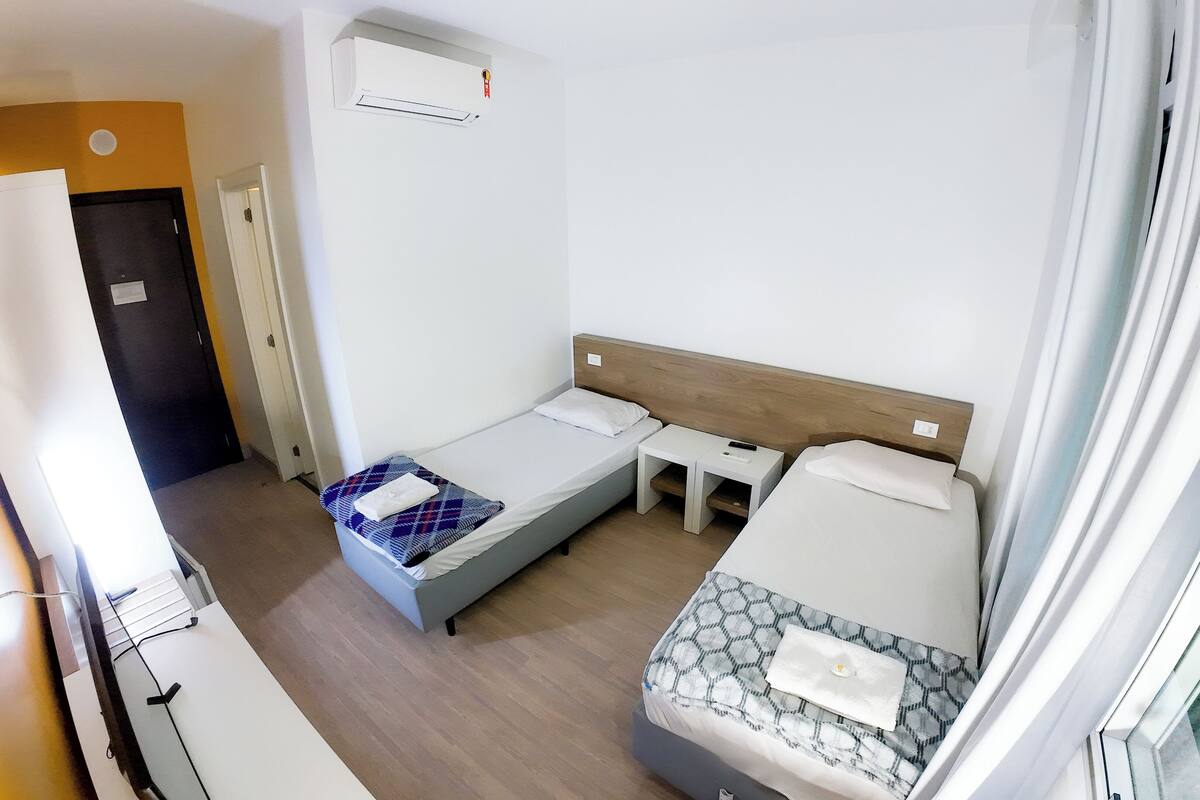Ein kleines Hotelzimmer mit zwei Einzelbetten, einem Tisch, einem Stuhl und einem fensterbepflanzten Fenster.