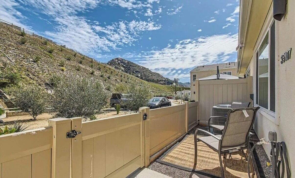 A balcony with a view of a hilly landscape, a beige fence, and a small patio area with chairs and a table.