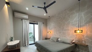A bedroom with a bed, a ceiling fan, a nightstand, an ironing board, and a stone wall. - Rooms Zicatela (Puerto Escondido)