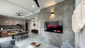 A modern living room with a flat-screen TV mounted on the wall, a dining area with a wooden table and chairs, and a kitchen in the background. - Rooms Zicatela (Puerto Escondido)
