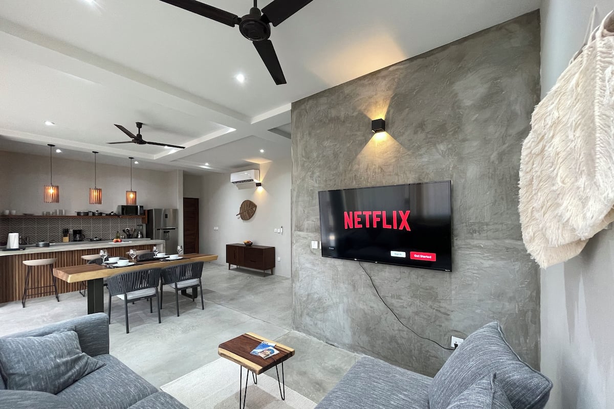 A modern living room with a flat-screen TV mounted on the wall, a dining area with a wooden table and chairs, and a kitchen in the background.