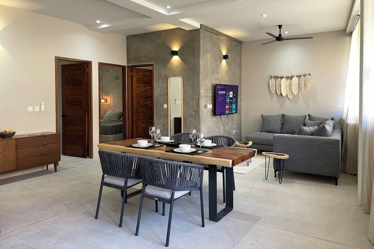 A modern living room with a dining area, grey sofa, wooden dining table, and ceiling fans.