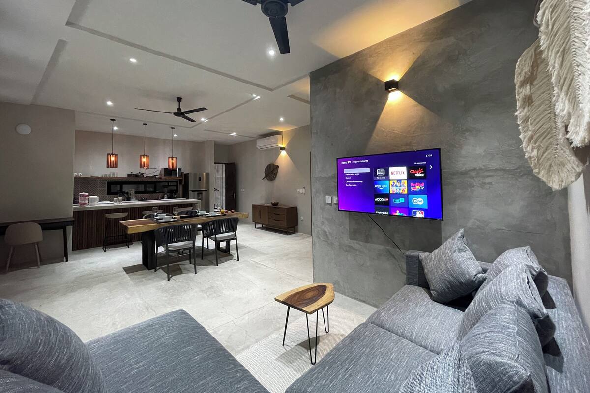 A modern living room with a grey sofa, a wooden coffee table, and a flat-screen TV mounted on the wall. There is a ceiling fan, recessed lighting, and a kitchen area in the background.