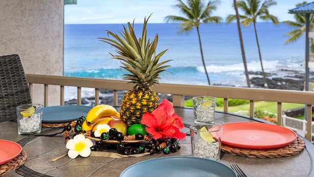 A table set for a meal with tropical fruits, a red flower, and a view of the ocean.
