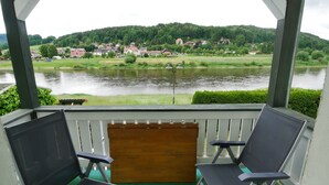 Un balcon avec deux chaises et une table, donnant sur une rivière et un village.