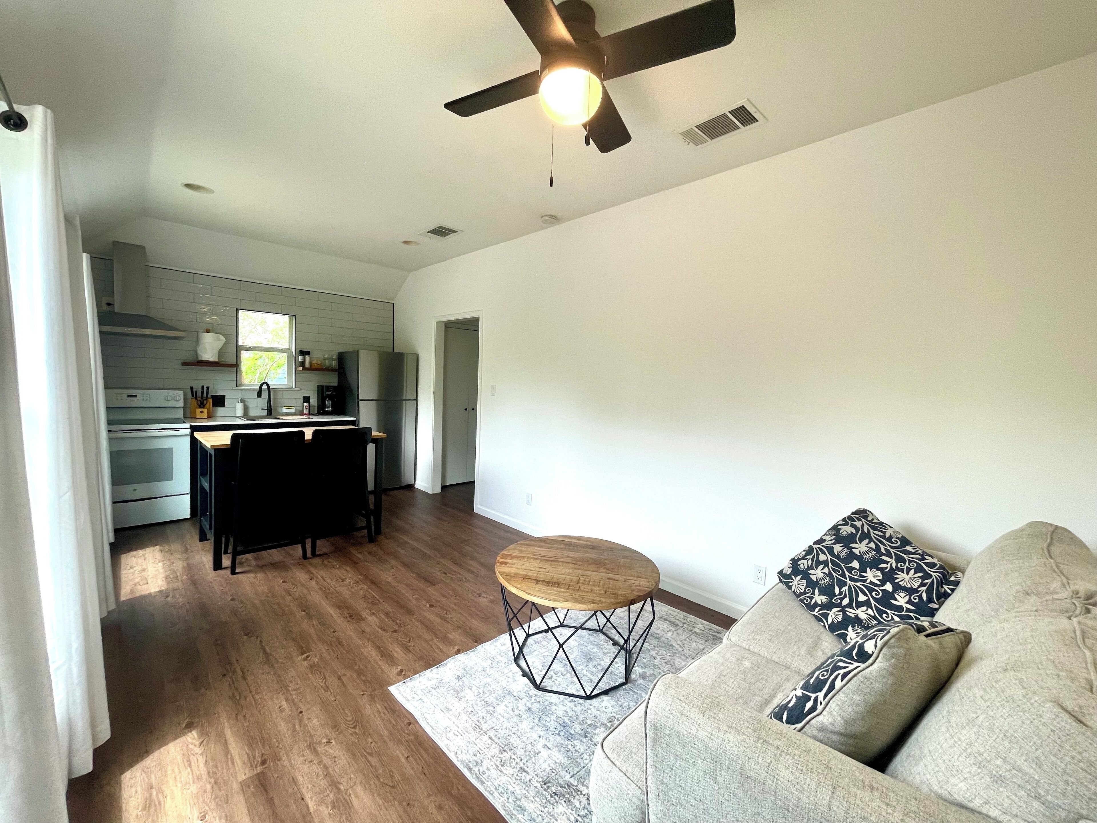 A modern living room with a wooden floor, a round coffee table, and a sofa with patterned cushions. A kitchen with white cabinets and a built-in oven is visible in the background.