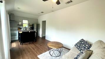 A modern living room with a wooden floor, a round coffee table, and a sofa with patterned cushions. A kitchen with white cabinets and a built-in oven is visible in the background.