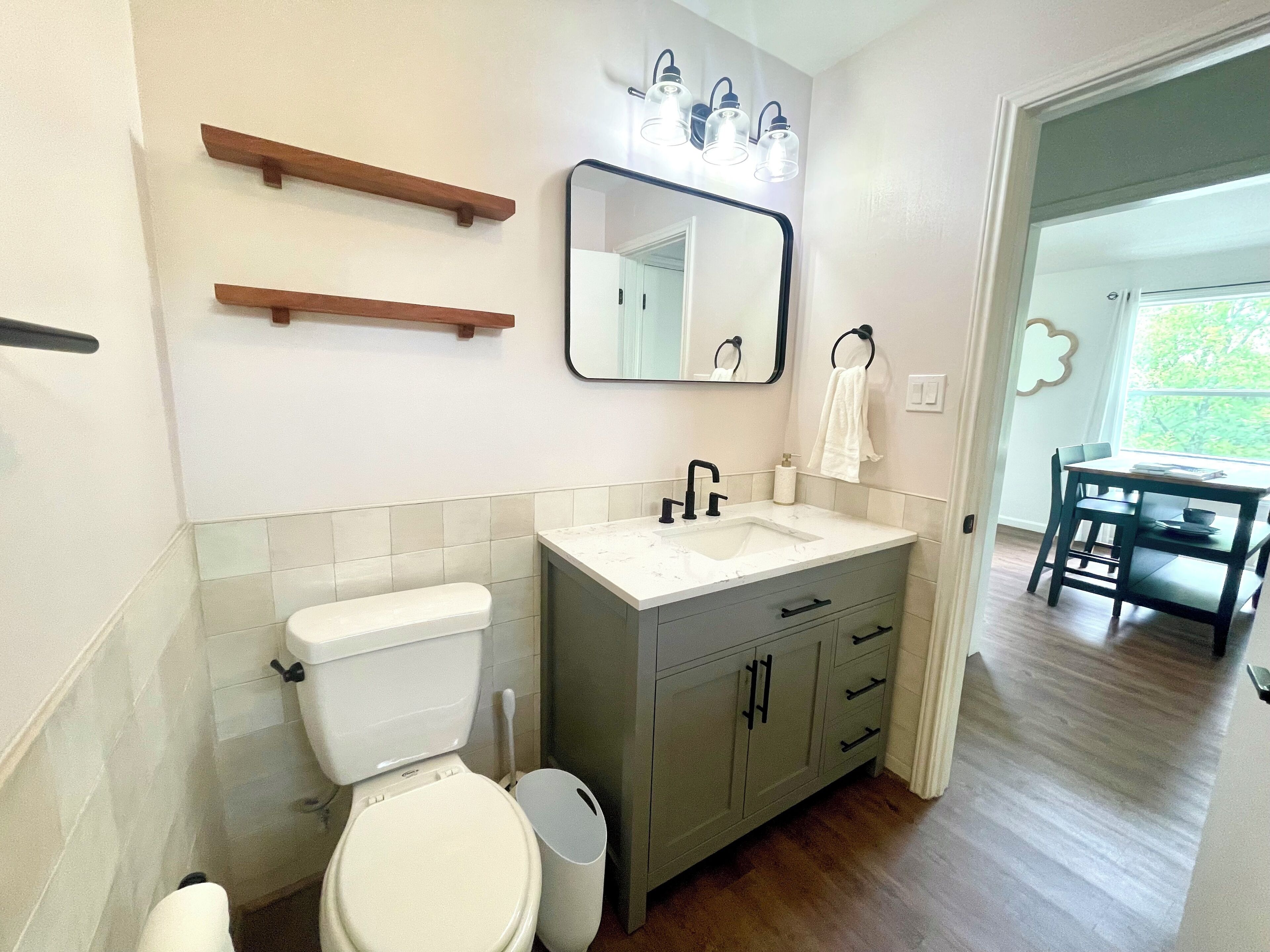 Een badkamer met toilet, wastafel, badkamermeubel en spiegel. Boven de wastafel zijn houten planken en een raam met uitzicht op groen.