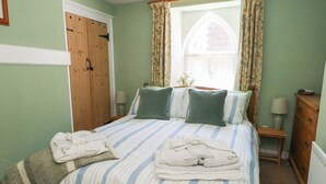 Ein ordentlich gemachtes Bett mit gestreifter Bettwäsche und Kissen, ein Holzschrank, ein Fenster mit blumigen Vorhängen und ein Nachttisch mit einer Lampe.