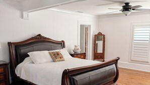Ein Schlafzimmer mit einem großen Bett, einem Holzkopfende, einem Deckenventilator, einer Holzkommode, einem Spiegel und einem Fenster mit Jalousien.