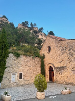 A stone building with a wooden door and a cross on top, surrounded by greenery and cobblestone pavement. - Marinette's house (Séguret)