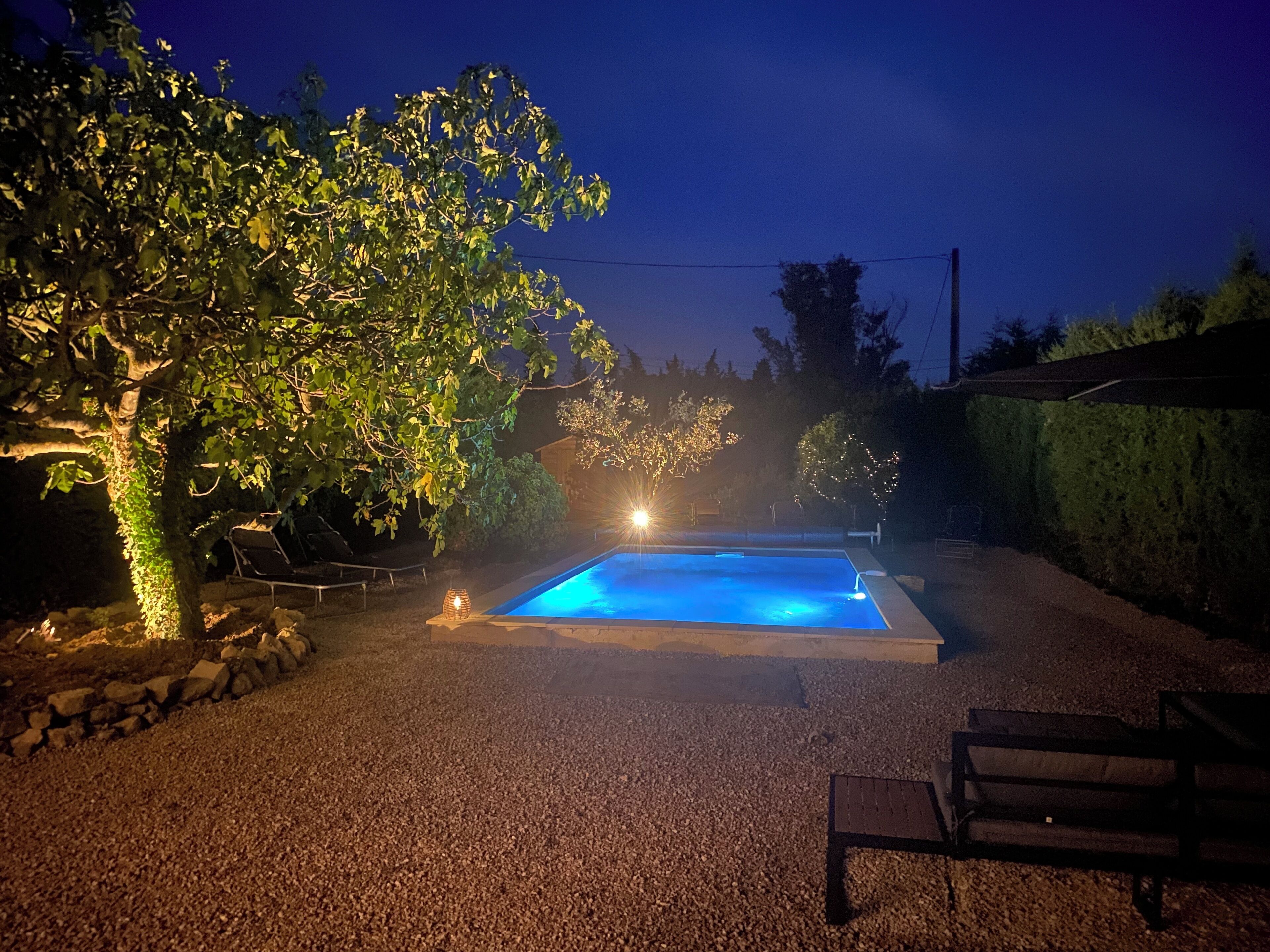 A swimming pool with a stone border, surrounded by gravel, trees, and a bench.