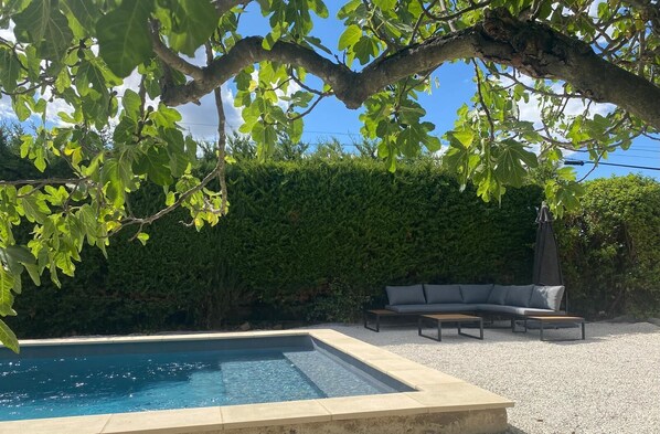 A swimming pool with a stone edge, surrounded by a gravel area. There is a gray sofa and two wooden tables. - Marinette's house (Séguret)
