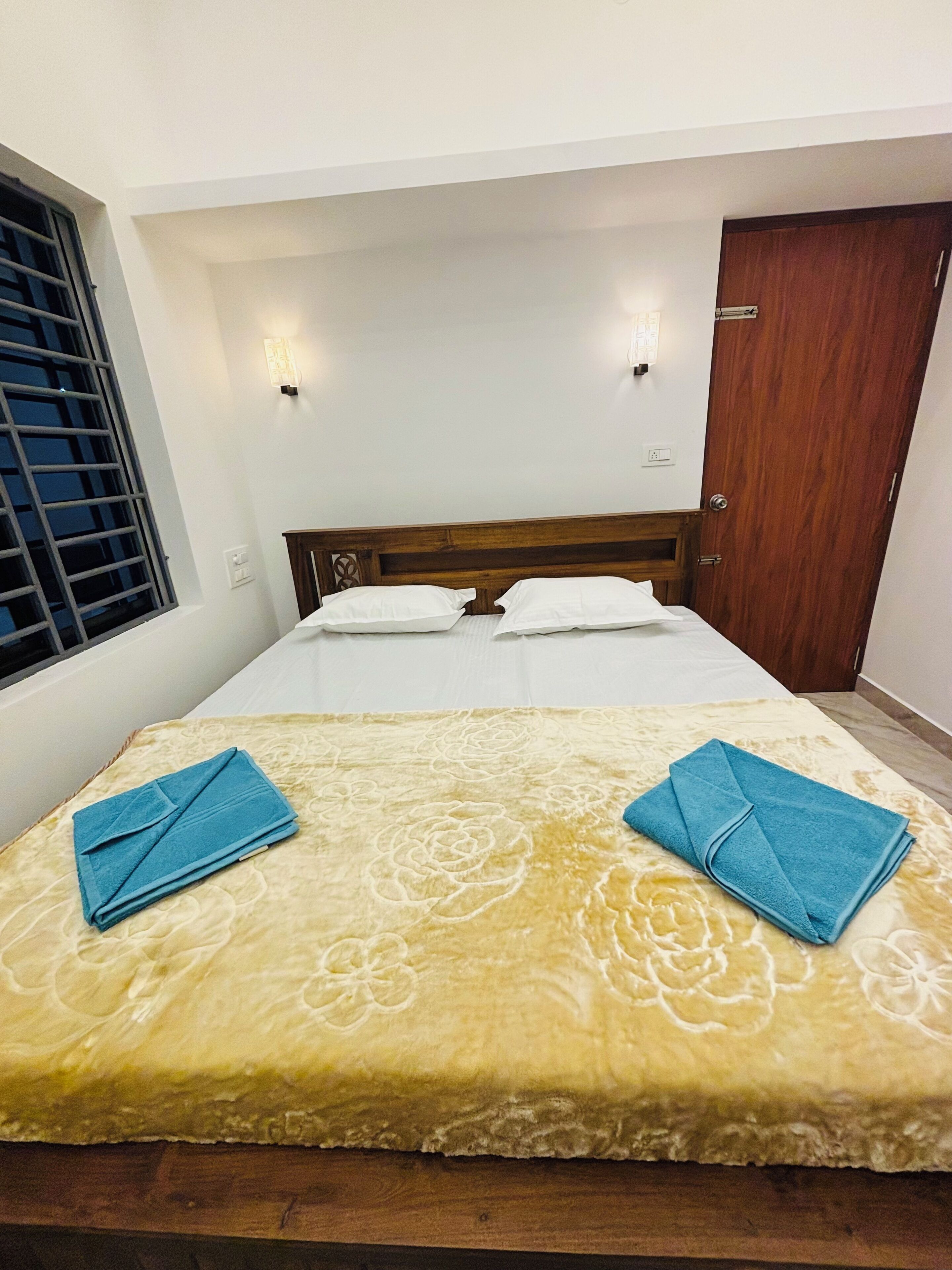 A hotel room with a bed, two pillows, a wooden headboard, a window with blinds, and a door.