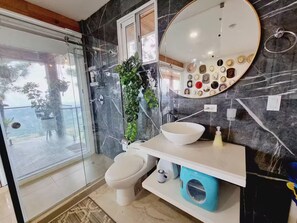 A bathroom with a glass-enclosed shower, a toilet, a sink with a white basin, a blue laundry basket, and a mirror with decorative items. - Cabaña Jacuzzi Paisaje Vista Increible Altos Maria (Sorá)