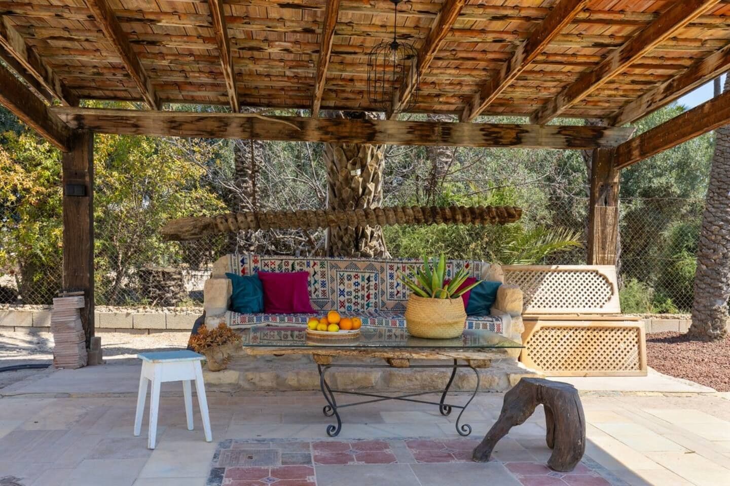 An outdoor seating area with a wooden pergola, a patterned bench, and a table with fruits and a potted plant.