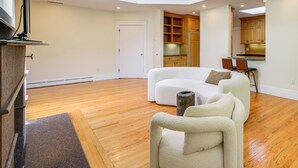 A spacious living area with a curved white sofa, a wooden dining table, and a kitchen with wooden cabinets. - 18 Mi to Boston - Stylish Unit in Upscale Enclave! (Weston)
