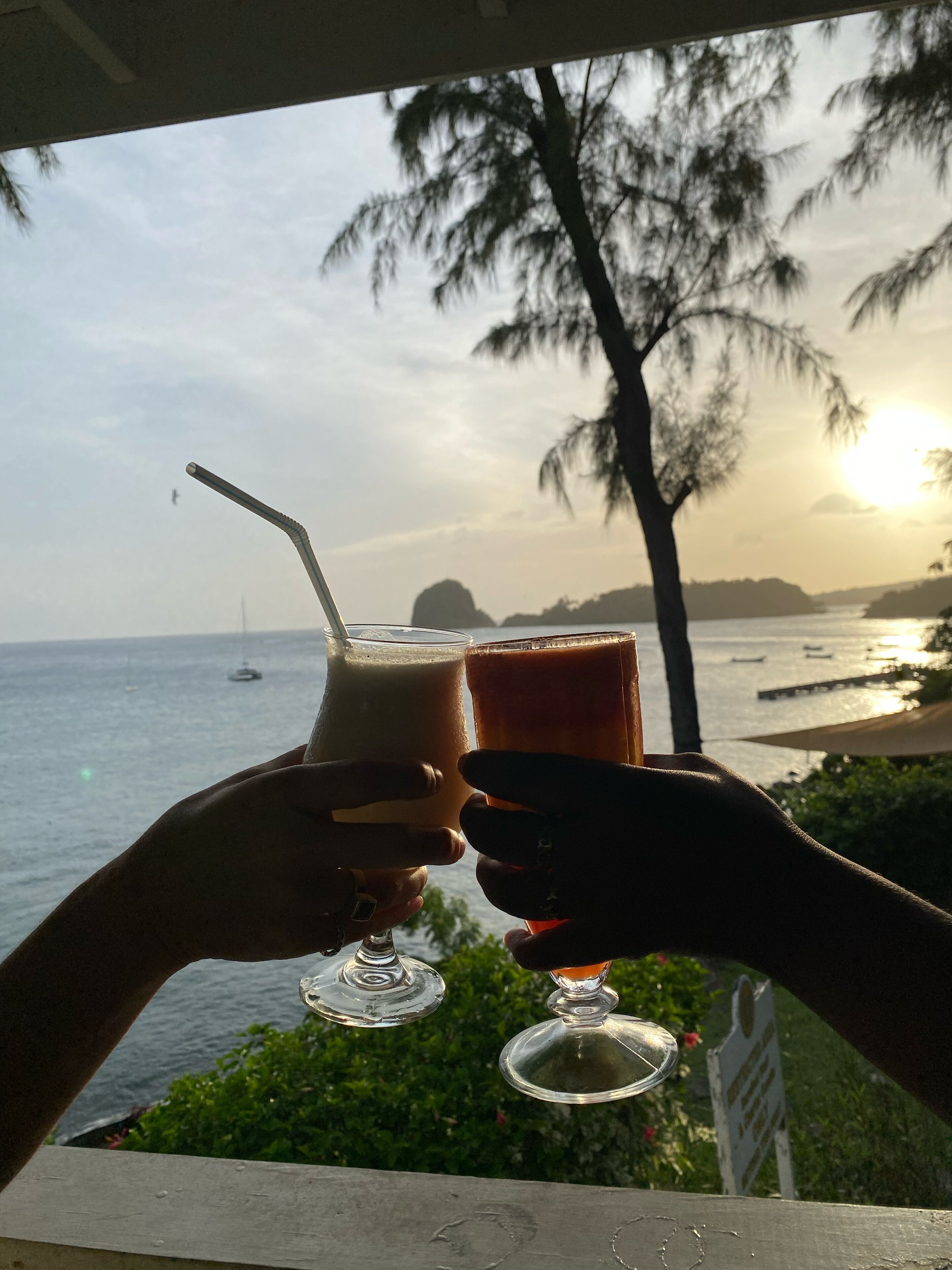Dos manos sosteniendo vasos con bebidas, con vistas a la costa al atardecer, palmeras y un barco a lo lejos.