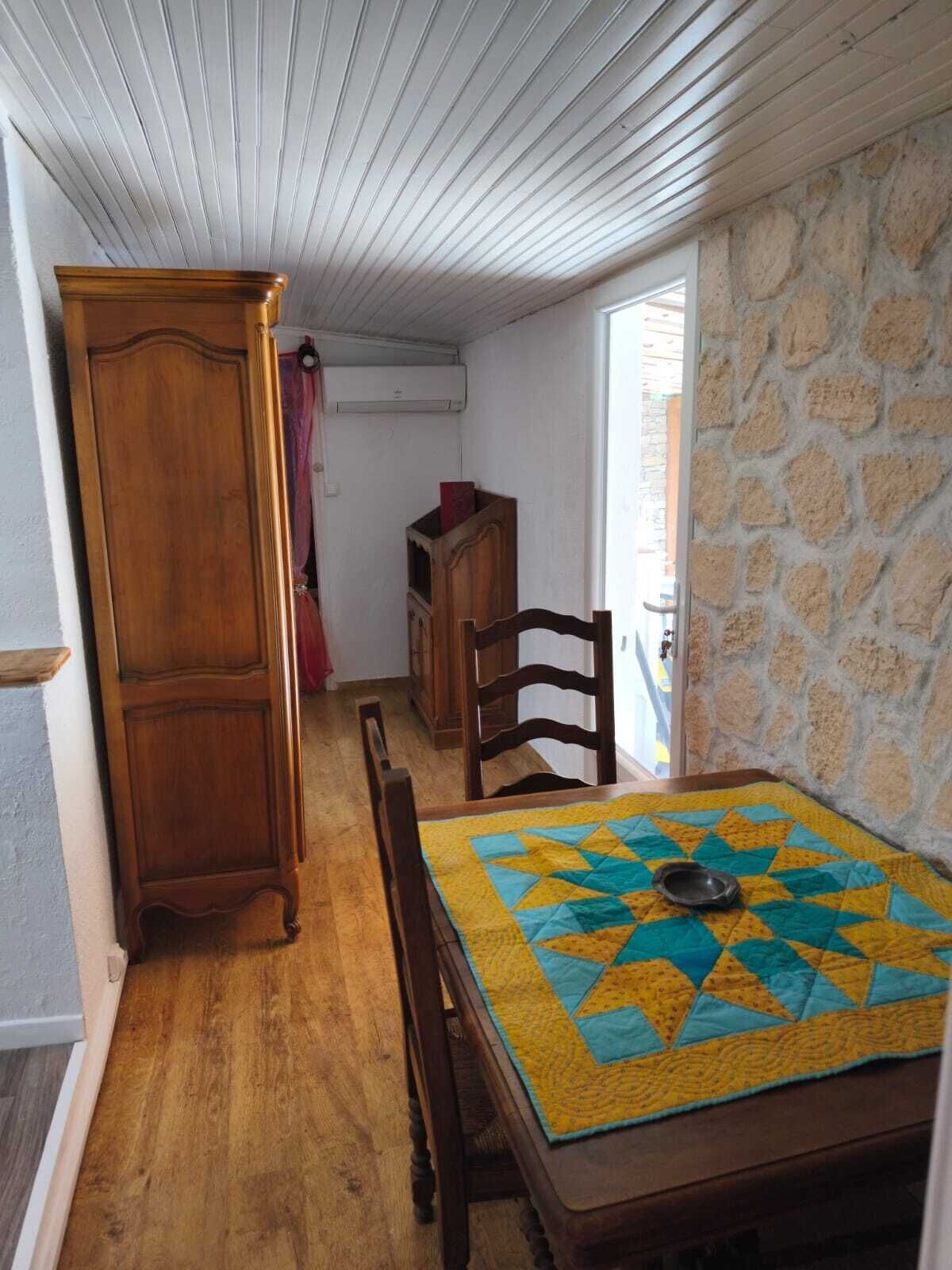 A dining area with a wooden table, chairs, and a quilted table runner. A wooden cabinet and a small wooden shelf are visible in the background.